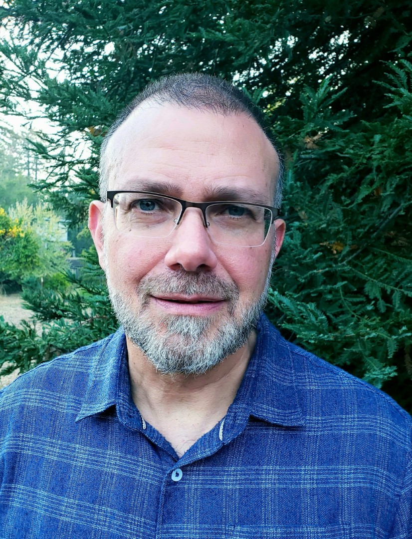 A man with glasses and a beard standing in front of trees.